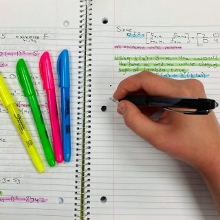 A hand writing notes in a notebook beside colourful highlighters