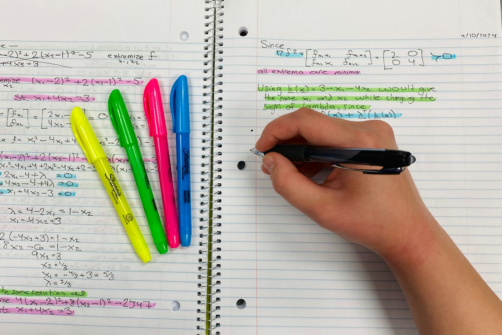 A hand writing notes in a notebook beside colourful highlighters