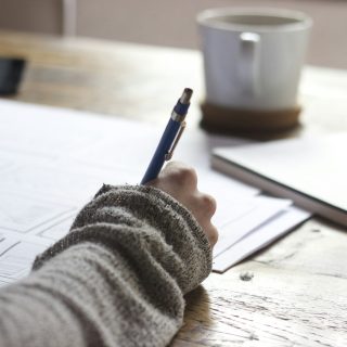 A person writing notes at a wooden table beside a mug