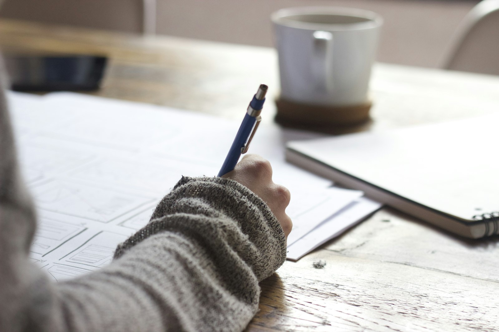 A person writing notes at a wooden table beside a mug